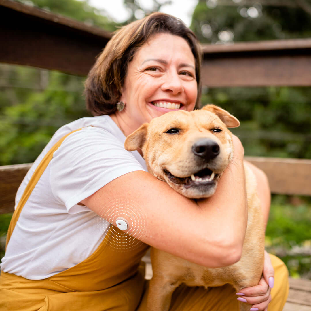Woman hugging a smiling dog outdoors, showcasing a continuous glucose monitoring system on her arm.